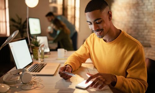 feliz-empresario-afro-americano-usando-telefone-celular-e-cartao-de-credito-de-pagamentos-online-enquanto-trabalhava-a-noite-no-escritorio